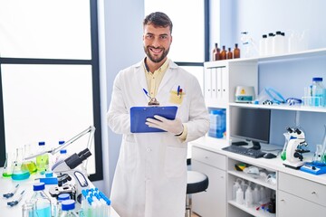 Naklejka premium Young hispanic man wearing scientist uniform writing on clipboard at laboratory