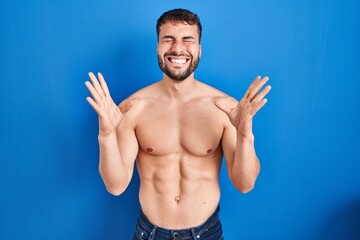 Handsome hispanic man standing shirtless celebrating mad and crazy for success with arms raised and...