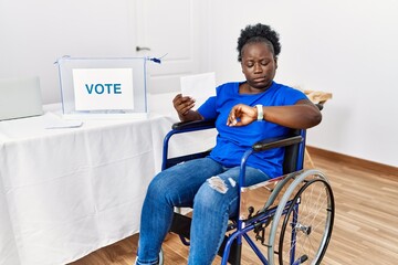 Young african woman sitting on wheelchair voting putting envelop in ballot box checking the time on...