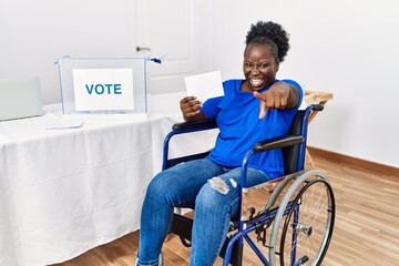 Young african woman sitting on wheelchair voting putting envelop in ballot box pointing to you and...
