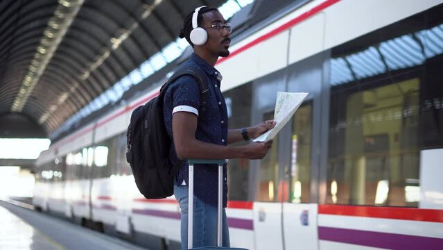 African male looking at his map while a train pass by. He is holding his suitcase at a train station