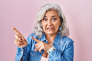 Middle age woman with grey hair standing over pink background pointing aside worried and nervous...
