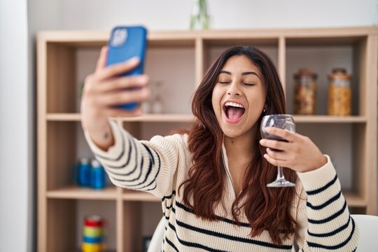Hispanic young business woman taking a selfie picture drinking a glass of wine smiling and laughing hard out loud because funny crazy joke.
