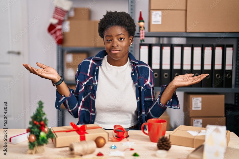 Sticker African american woman working at small business doing christmas decoration clueless and confused expression with arms and hands raised. doubt concept.