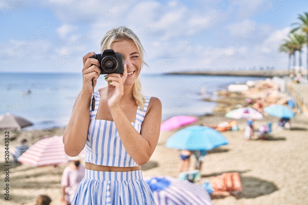 Poster young blonde girl smiling happy using camera at the beach.