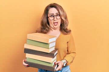 Middle age caucasian woman holding a pile of books in shock face, looking skeptical and sarcastic, surprised with open mouth