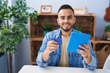 Young hispanic man using touchpad and credit card sitting on table at home