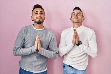 Homosexual couple standing over pink background begging and praying with hands together with hope...