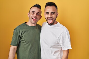 Homosexual couple standing over yellow background sticking tongue out happy with funny expression. emotion concept.