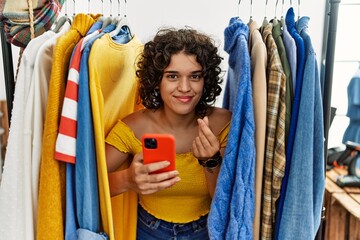Young hispanic woman searching clothes on clothing rack using smartphone doing money gesture with...