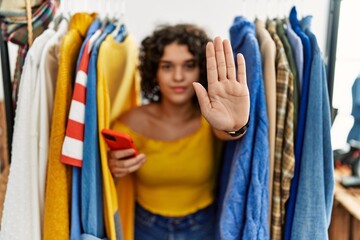 Young hispanic woman searching clothes on clothing rack using smartphone doing stop sing with palm...