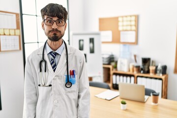 Hispanic man with beard wearing doctor uniform and stethoscope at the office puffing cheeks with funny face. mouth inflated with air, crazy expression. - obrazy, fototapety, plakaty