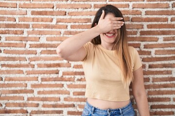 Young brunette woman standing over bricks wall smiling and laughing with hand on face covering eyes for surprise. blind concept.