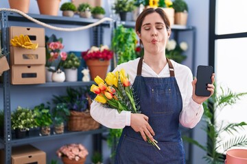 Brunette woman working at florist shop holding smartphone making fish face with mouth and squinting eyes, crazy and comical.