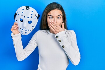 Young caucasian girl holding hockey mask covering mouth with hand, shocked and afraid for mistake....