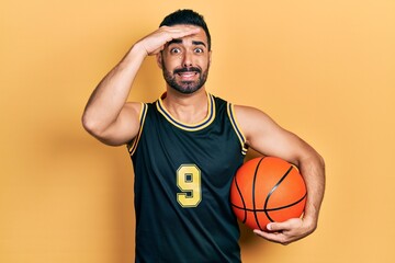 Handsome hispanic man with beard holding basketball ball stressed and frustrated with hand on head, surprised and angry face