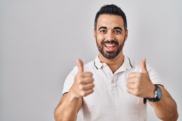 Young hispanic man with beard wearing casual clothes over white background success sign doing positive gesture with hand, thumbs up smiling and happy. cheerful expression and winner gesture.