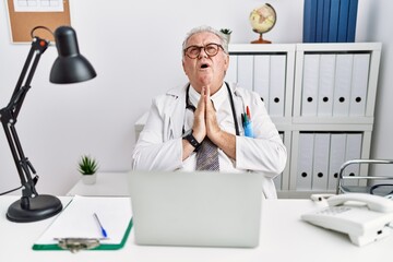 Senior caucasian man wearing doctor uniform and stethoscope at the clinic begging and praying with hands together with hope expression on face very emotional and worried. begging.