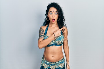 Young woman wearing bindi and traditional belly dance clothes surprised pointing with finger to the side, open mouth amazed expression.