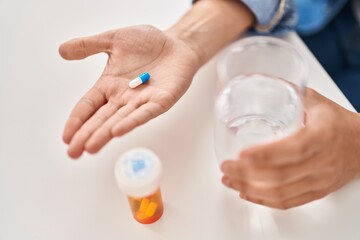 Young non binary man holding pill sitting on table at home