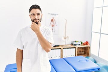 Fototapeta premium Young handsome man with beard working at pain recovery clinic looking confident at the camera with smile with crossed arms and hand raised on chin. thinking positive.