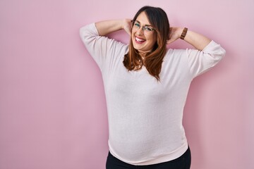 Pregnant woman standing over pink background relaxing and stretching, arms and hands behind head and neck smiling happy