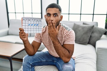 Young indian man holding travel calendar covering mouth with hand, shocked and afraid for mistake. surprised expression