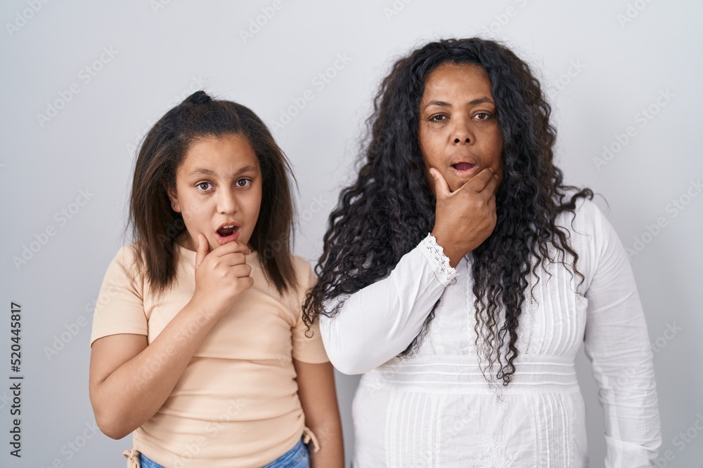 Sticker mother and young daughter standing over white background looking fascinated with disbelief, surprise