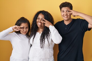 Obraz premium Family of mother, daughter and son standing over yellow background doing peace symbol with fingers over face, smiling cheerful showing victory