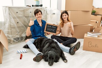 Young caucasian couple with dog holding our first home blackboard at new house doing happy thumbs up gesture with hand. approving expression looking at the camera showing success.