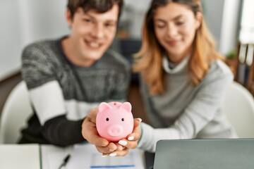 Young caucasian couple smiling happy holding piggy bank sitting on the table at home.
