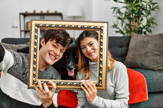 Young caucasian couple with dog making family picture holding empty frame at home.