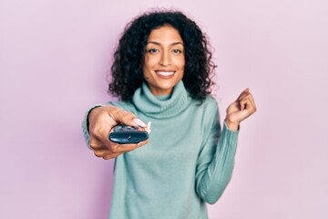 Young latin girl holding television remote control screaming proud, celebrating victory and success very excited with raised arm