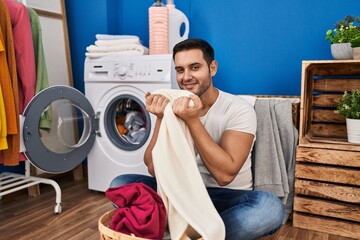 Young hispanic man smiling confident smelling clothes at laundry room