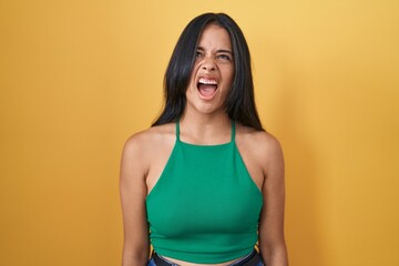 Brunette woman standing over yellow background angry and mad screaming frustrated and furious,...