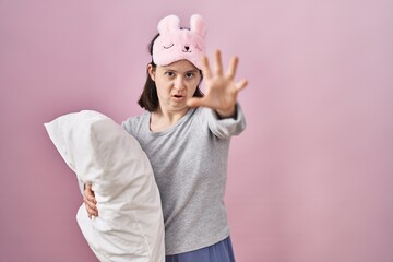 Woman with down syndrome wearing sleeping mask hugging pillow doing stop gesture with hands palms, angry and frustration expression