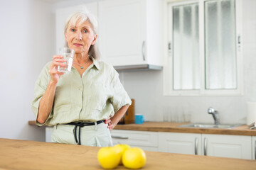 Cheerful senior woman standing in kitchen at home and drinking water.