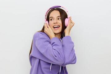 a happy woman stands on a white background in a purple tracksuit and pink headphones on her head, pressing them with both hands, looking away joyfully