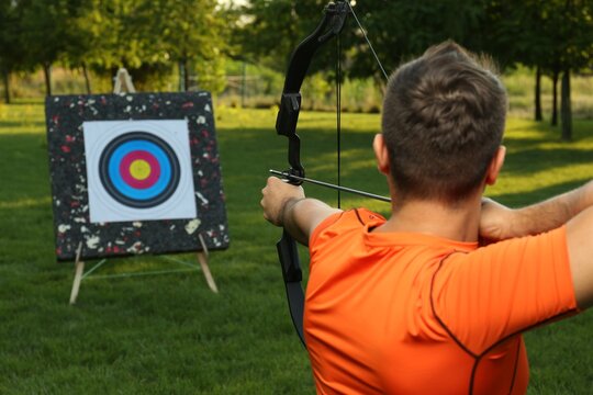 Man with bow and arrow aiming at archery target in park, back view