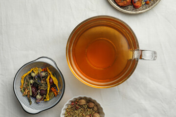 Cup of freshly brewed tea, dried herbs and berries on white fabric, flat lay