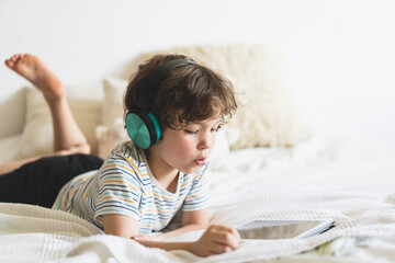 Cute boy lying on the bed at home and playing with pc tablet or reading online and listening to...