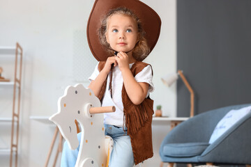 Cute little girl in hat and vest playing with rocking horse at home