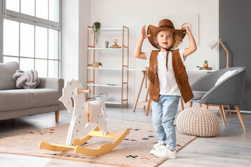 Funny little girl in hat and vest playing with rocking horse at home