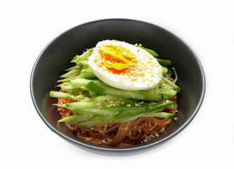 Korean cold noodles with spicy sauce, topped with cucumber, boiled egg and crushed sesame seeds in a black bowl. Bibim Guksu or Korean cold noodle salad (Spicy mixed noodles) Usually eat in Summer.