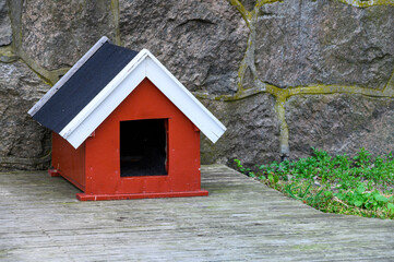 Fototapeta na wymiar Cute red doghouse with white roof trim on a weathered wood deck against a stone wall 