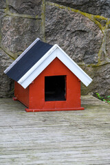 Fototapeta na wymiar Cute red doghouse with white roof trim on a weathered wood deck against a stone wall 