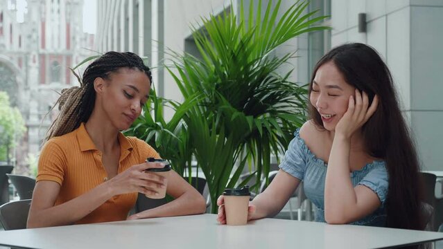 Young ebony mixed race girl with dreadlocks and attractive asian girl sit at a table and have cute conversation outdoors. Two young women talking and drinking coffee sitting in cafe.