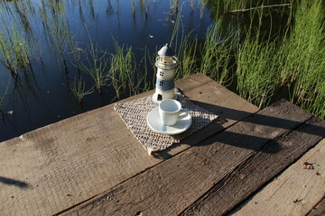 Obraz na płótnie Canvas Lighthouse on wooden pier on riverside, empty white cup and soucer, forest flowers in sunny summer morning