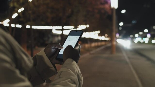 Unrecognizable Black man holding smartphone using taxi app to request ride late at night, selective focus shot