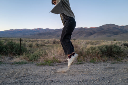 Short haired non-binary person jumps playfully on dusty road in open road landscape with mountains